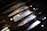 A set of Japanese chef knives with patterned blades is displayed in a wooden box. The blades show intricate patterns, possibly indicating Damascus steel. The knives have dark handles and are arranged neatly side by side. Reflections of lights can be seen on the glass covering.