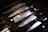A set of Japanese chef knives with patterned blades is displayed in a wooden box. The blades show intricate patterns, possibly indicating Damascus steel. The knives have dark handles and are arranged neatly side by side. Reflections of lights can be seen on the glass covering.