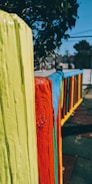A colorful wooden fence with brightly painted vertical planks in yellow, red, blue, and green. The fence is in an outdoor setting with trees in the background and sections of a playground or park visible.