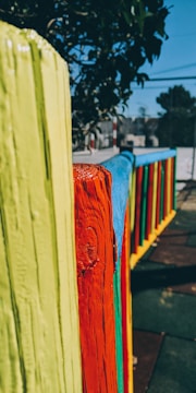 A colorful wooden fence with brightly painted vertical planks in yellow, red, blue, and green. The fence is in an outdoor setting with trees in the background and sections of a playground or park visible.