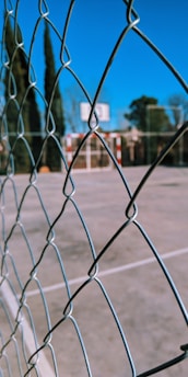 A chain-link fence in the foreground with a blurred sports field and goalpost in the background. Tall trees are visible, and the sky is clear and blue.