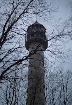 A tall tower with a cylindrical structure is surrounded by bare trees. The sky is overcast, and raindrops are visible against the dim lighting. Red lights are visible on the top part of the tower, adding an eerie atmosphere.