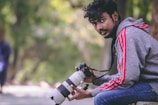 A person wearing a gray hoodie with red stripes and blue jeans is sitting outdoors holding a professional camera with a large lens. The background features blurred greenery, suggesting a park or natural setting. The individual's expression appears focused and relaxed.