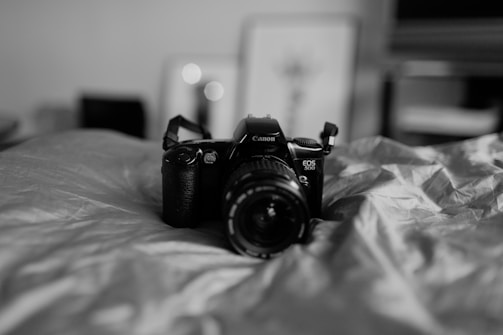 A black and white photograph featuring a Canon EOS 500 camera resting on a crumpled fabric surface. The background is softly out of focus, creating a dreamy atmosphere with some abstract shapes and frames visible, enhancing the depth of field.