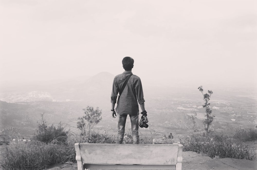 grayscale photography of man standing on cliff near bench holding DSLR camera