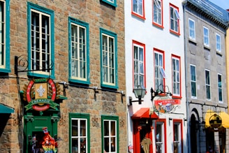 A street view of colorful historical buildings with stone and painted facades, featuring signage for souvenir shops and a hotel. The windows are framed in contrasting vibrant colors, and there are decorative elements and hanging signs.