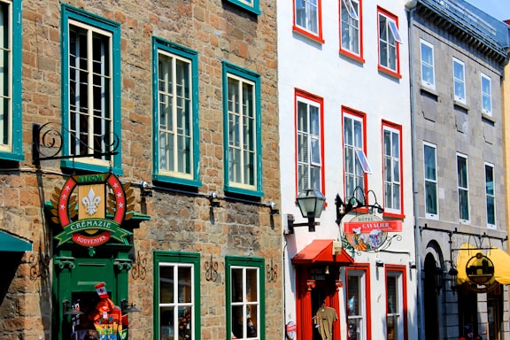 A street view of colorful historical buildings with stone and painted facades, featuring signage for souvenir shops and a hotel. The windows are framed in contrasting vibrant colors, and there are decorative elements and hanging signs.