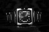 A sleek, metallic wristwatch with a square face and bold numerals displayed prominently. The watch features a sturdy metal band and a minimalist design, set against a dark, textured background.