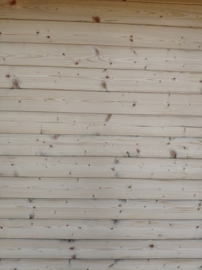 A series of horizontal wooden planks with visible knots and grain patterns. The wood appears freshly cut or possibly sanded, showcasing a natural light brown color.