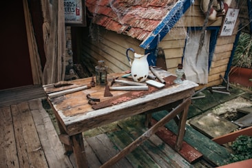 An old wooden table is cluttered with various rustic tools and objects, including a worn-out saw, a lantern, and a rusted pitcher. The setting is outdoors next to a quaint, weathered wooden building with a sign that reads 'No Rooms for Rent'. The roof is covered in distressed red shingles, and cobwebs are visible throughout the scene.