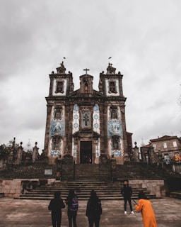 An ornate historical church with blue and white tile decorations stands prominently under a cloudy sky. Stone steps lead up to the entrance, where several people are gathered, one of whom is wearing a bright orange coat.