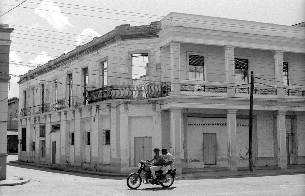 建物を通り過ぎるオートバイに乗る2人の男のグレースケール写真