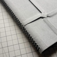 A close-up view of a textured gray fabric with black stitching along the edge, resting on a grid-patterned surface. The fabric fold is secured with a square knot.