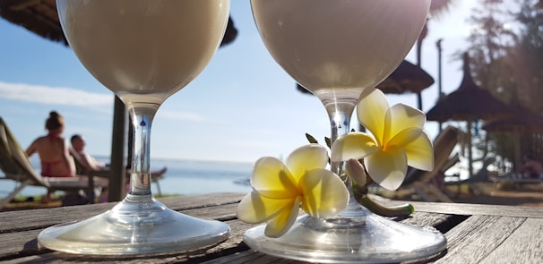 Two frothy drinks in stemmed glasses placed on a wooden surface with tropical flowers beside them. The background displays a beach scene with straw-roofed umbrellas and people relaxing on lounge chairs, set against a clear blue sky and ocean view.