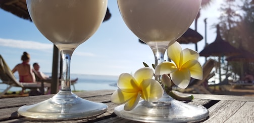 Two frothy drinks in stemmed glasses placed on a wooden surface with tropical flowers beside them. The background displays a beach scene with straw-roofed umbrellas and people relaxing on lounge chairs, set against a clear blue sky and ocean view.