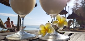 Two frothy drinks in stemmed glasses placed on a wooden surface with tropical flowers beside them. The background displays a beach scene with straw-roofed umbrellas and people relaxing on lounge chairs, set against a clear blue sky and ocean view.
