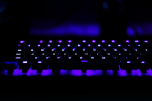 A computer keyboard illuminated with vibrant purple lights creates a striking visual against a dark background. The keys are distinctly visible with the LED backlighting, adding a modern and futuristic aesthetic.