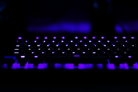 A computer keyboard illuminated with vibrant purple lights creates a striking visual against a dark background. The keys are distinctly visible with the LED backlighting, adding a modern and futuristic aesthetic.