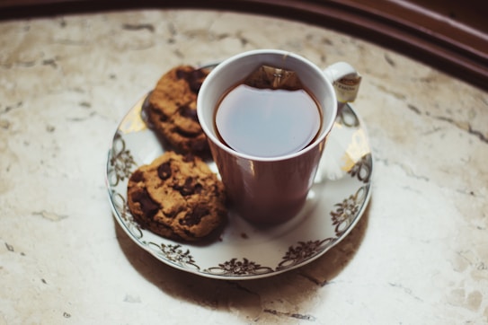 A cup of tea with a teabag inside sits on an ornate saucer next to two chocolate chip cookies. The surface beneath is a textured stone or marble-like countertop.