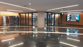 A modern indoor space with polished dark marble floors, bright linear ceiling lights, and wooden paneling along the walls. The area features glass doors with signage for 'The Wash' and a digital display on the wall showing multiple data screens.