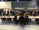 A group of people are gathered in a modern conference or meeting space with tables, chairs, and plants. Individuals are engaged in conversations and interactions, suggesting a professional or networking event. The space is well-lit with hanging lights above the tables and a dark gray background wall featuring signs or logos.