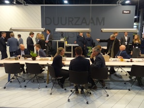 A group of people are gathered in a modern conference or meeting space with tables, chairs, and plants. Individuals are engaged in conversations and interactions, suggesting a professional or networking event. The space is well-lit with hanging lights above the tables and a dark gray background wall featuring signs or logos.