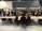 A group of people are gathered in a modern conference or meeting space with tables, chairs, and plants. Individuals are engaged in conversations and interactions, suggesting a professional or networking event. The space is well-lit with hanging lights above the tables and a dark gray background wall featuring signs or logos.
