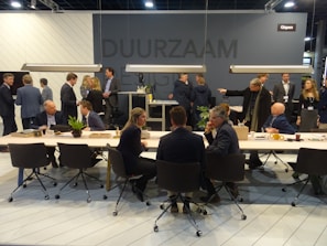 A group of people are gathered in a modern conference or meeting space with tables, chairs, and plants. Individuals are engaged in conversations and interactions, suggesting a professional or networking event. The space is well-lit with hanging lights above the tables and a dark gray background wall featuring signs or logos.