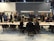 A group of people are gathered in a modern conference or meeting space with tables, chairs, and plants. Individuals are engaged in conversations and interactions, suggesting a professional or networking event. The space is well-lit with hanging lights above the tables and a dark gray background wall featuring signs or logos.