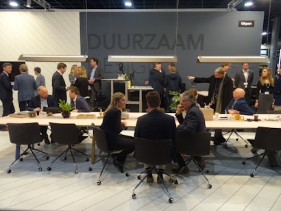 A group of people are gathered in a modern conference or meeting space with tables, chairs, and plants. Individuals are engaged in conversations and interactions, suggesting a professional or networking event. The space is well-lit with hanging lights above the tables and a dark gray background wall featuring signs or logos.