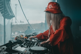 A DJ wearing a red outfit and bucket hat is performing at a mix deck. In the background, a person is visible near the window with a partially cloudy sky outside. The DJ equipment is adorned with flowers.
