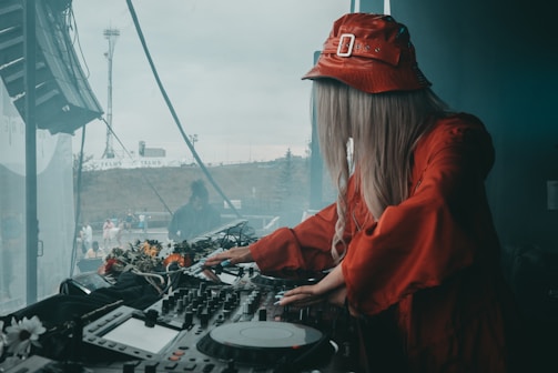 A DJ wearing a red outfit and bucket hat is performing at a mix deck. In the background, a person is visible near the window with a partially cloudy sky outside. The DJ equipment is adorned with flowers.