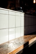 A wooden countertop with a glossy finish is illuminated by natural sunlight, casting a soft shadow on a backdrop of glossy white tiles arranged in a grid. A black coffee cup and saucer rest on the counter.