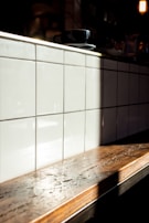 A wooden countertop with a glossy finish is illuminated by natural sunlight, casting a soft shadow on a backdrop of glossy white tiles arranged in a grid. A black coffee cup and saucer rest on the counter.