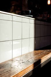 A wooden countertop with a glossy finish is illuminated by natural sunlight, casting a soft shadow on a backdrop of glossy white tiles arranged in a grid. A black coffee cup and saucer rest on the counter.