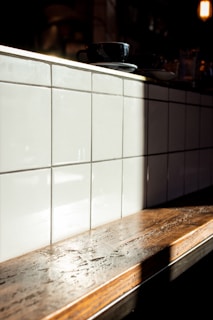 A wooden countertop with a glossy finish is illuminated by natural sunlight, casting a soft shadow on a backdrop of glossy white tiles arranged in a grid. A black coffee cup and saucer rest on the counter.
