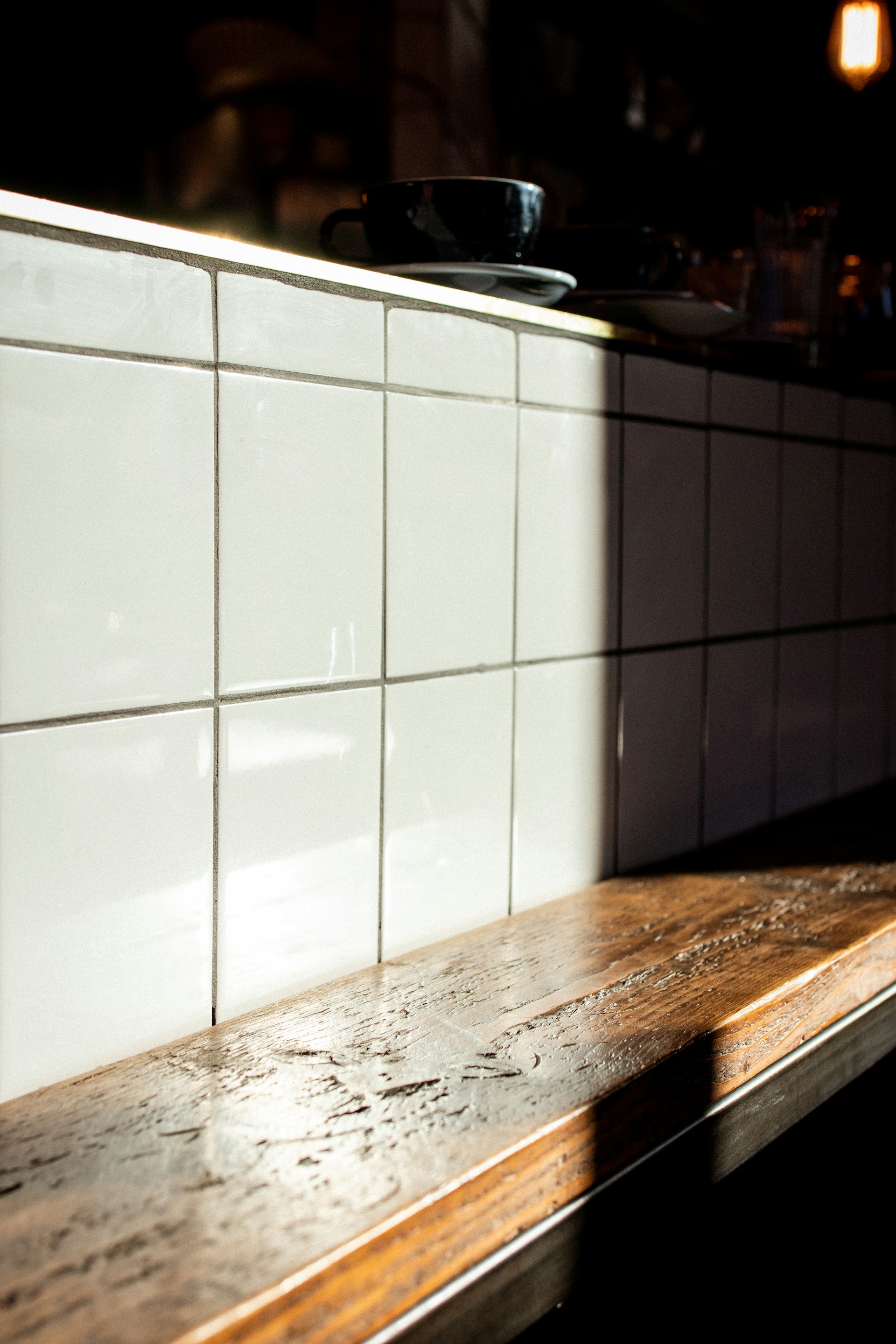 A wooden countertop with a glossy finish is illuminated by natural sunlight, casting a soft shadow on a backdrop of glossy white tiles arranged in a grid. A black coffee cup and saucer rest on the counter.