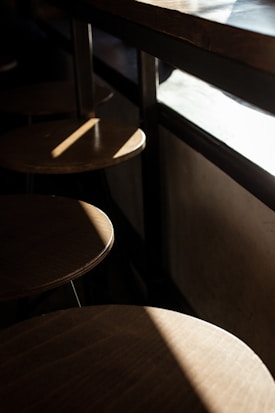 A series of wooden stools line up next to a counter, with sunlight streaming in from the window, casting distinct shadows and creating a warm, cozy atmosphere.