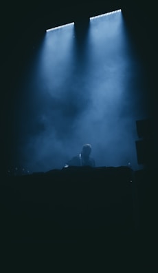 A person is positioned behind a DJ booth, enveloped in dim lighting and mist. Overhead, bright spotlights cast beams of light, creating a dramatic ambiance with shadows and highlights.