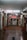 A red car is suspended on a vehicle lift in the middle of an auto repair garage. The garage has a high ceiling, fluorescent lights, and appears to be equipped with various tools and machinery along the walls. The floor of the garage is slightly wet and has a drain in the middle. There is a sign in Italian visible on the right side of the image.