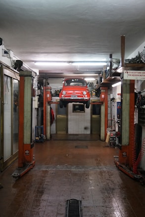 A red car is suspended on a vehicle lift in the middle of an auto repair garage. The garage has a high ceiling, fluorescent lights, and appears to be equipped with various tools and machinery along the walls. The floor of the garage is slightly wet and has a drain in the middle. There is a sign in Italian visible on the right side of the image.