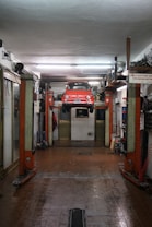 A red car is suspended on a vehicle lift in the middle of an auto repair garage. The garage has a high ceiling, fluorescent lights, and appears to be equipped with various tools and machinery along the walls. The floor of the garage is slightly wet and has a drain in the middle. There is a sign in Italian visible on the right side of the image.