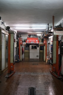 A red car is suspended on a vehicle lift in the middle of an auto repair garage. The garage has a high ceiling, fluorescent lights, and appears to be equipped with various tools and machinery along the walls. The floor of the garage is slightly wet and has a drain in the middle. There is a sign in Italian visible on the right side of the image.