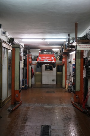 A red car is suspended on a vehicle lift in the middle of an auto repair garage. The garage has a high ceiling, fluorescent lights, and appears to be equipped with various tools and machinery along the walls. The floor of the garage is slightly wet and has a drain in the middle. There is a sign in Italian visible on the right side of the image.