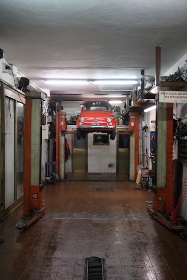 A red car is suspended on a vehicle lift in the middle of an auto repair garage. The garage has a high ceiling, fluorescent lights, and appears to be equipped with various tools and machinery along the walls. The floor of the garage is slightly wet and has a drain in the middle. There is a sign in Italian visible on the right side of the image.