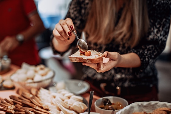 A person spreads a creamy topping on a slice of bread using a spoon. Various food items, possibly snacks or appetizers, are laid out on a table, including what appear to be dumplings and breadsticks. The scene suggests a casual food preparation or serving setting.