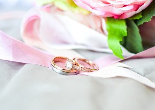 A close-up of two intertwined rings placed on a soft, pink ribbon. One ring is gold with a prominent diamond, while the other appears to be a simple silver band. In the background, there are hints of a pink and white flower with green leaves, adding a touch of elegance to the composition.