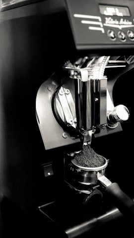 An espresso machine's portafilter is being filled with freshly ground coffee. The setup is sleek and modern, featuring a metallic finish with some controls visible at the top.