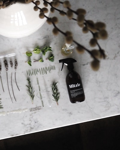 A variety of plant sprigs and leaves are neatly arranged on a light grey marble surface. A dark spray bottle labeled 'Mitzie' is positioned on the right side. There are also two small droppers and a small glass vial with liquid near the center. Blurred foliage appears at the top edges of the image.