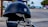 A black motorcycle helmet with a metallic emblem is placed on a parked motorcycle handlebar. The background features a coastal setting, including a road, a few parked cars, palm trees, and an ocean view further in the distance.